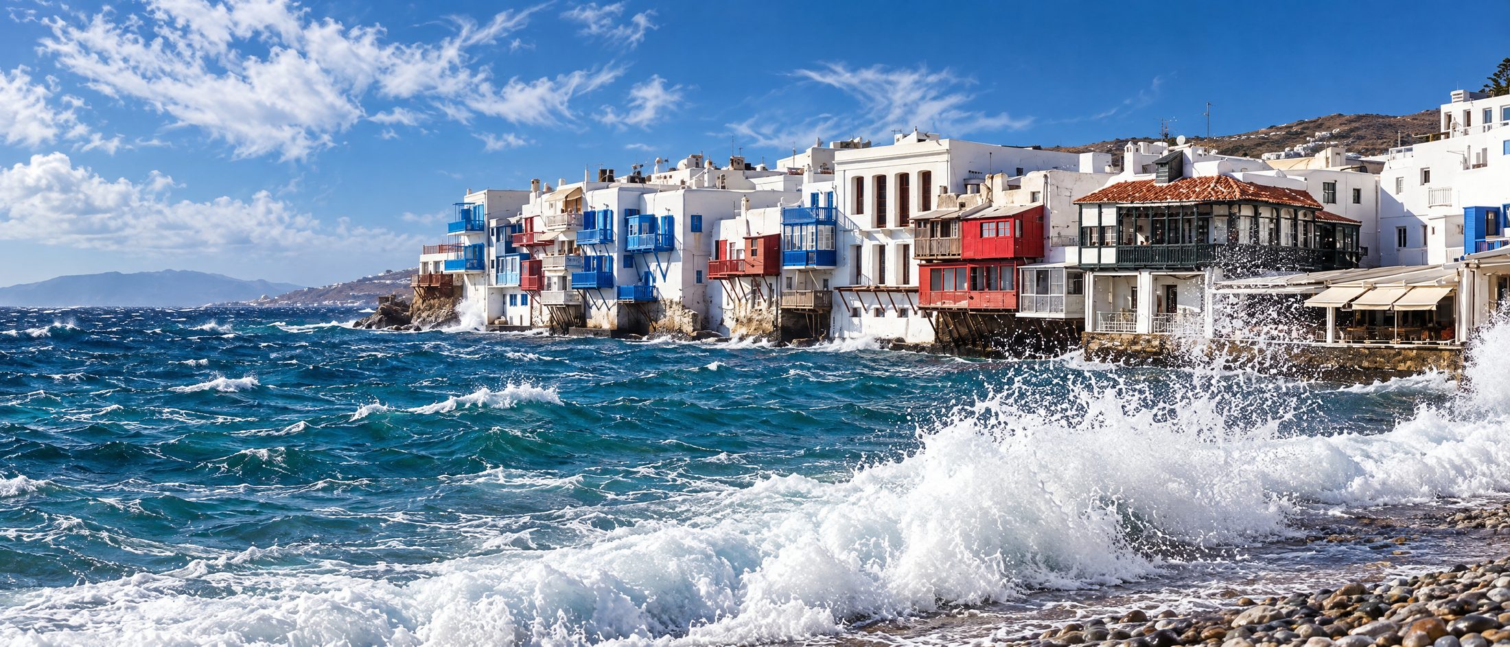A strong meltemi running past the Mykonos waterfront, whitecaps across the bay and spray against the houses