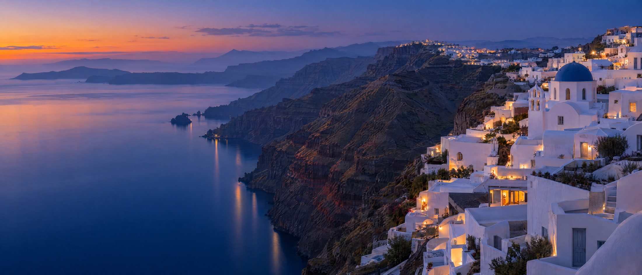 Whitewashed Cycladic houses and a blue-domed chapel on a caldera cliff at golden hour