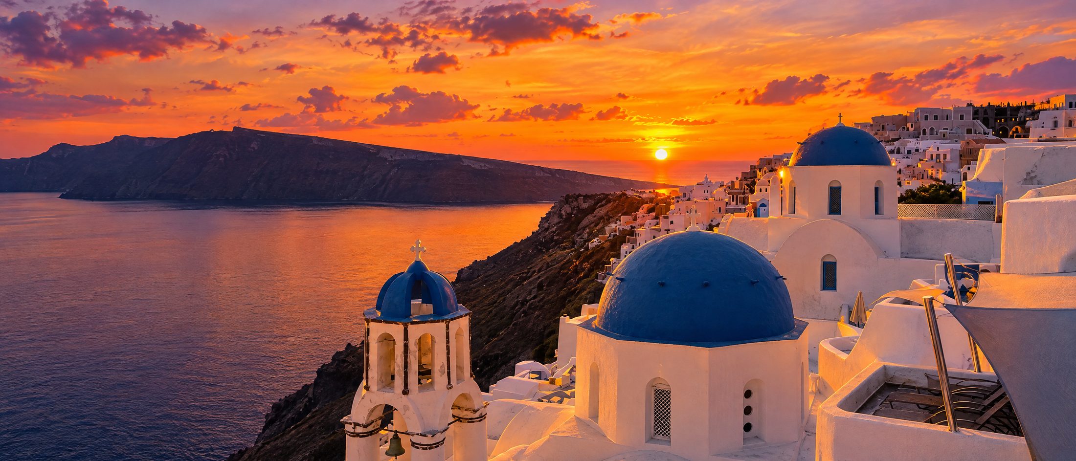 A blue-domed church on the Santorini caldera rim with the sun setting over the flooded caldera